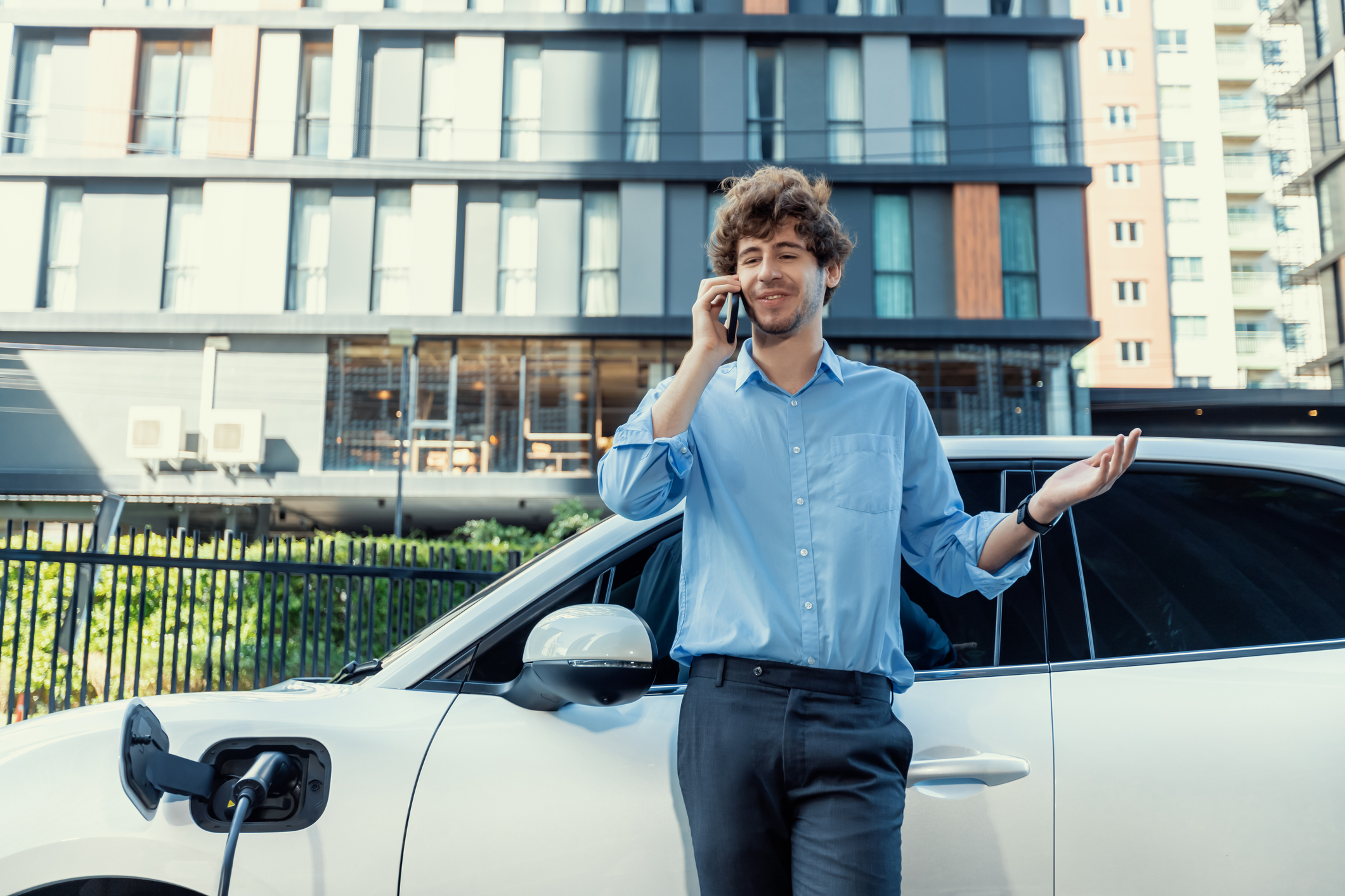 Progressive businessman talking on the phone, leaning on electric car recharging with public EV charging station, apartment condo residential building on the background as green city lifestyle.